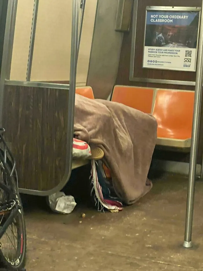 Person covered with a blanket lying on the floor of a NYC subway car next to empty orange seats and a bicycle.