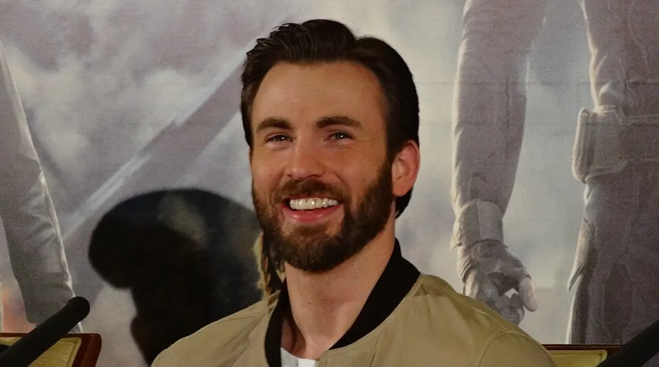 Chris Evans smiling at a press event, wearing a beige jacket with a beard and styled hair.