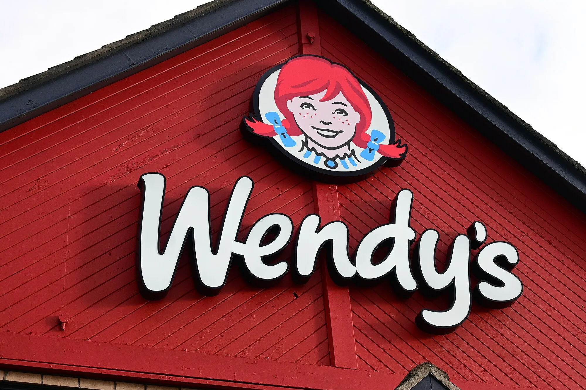 Wendy's logo featuring the red-haired girl on a red wooden building exterior under a cloudy sky.