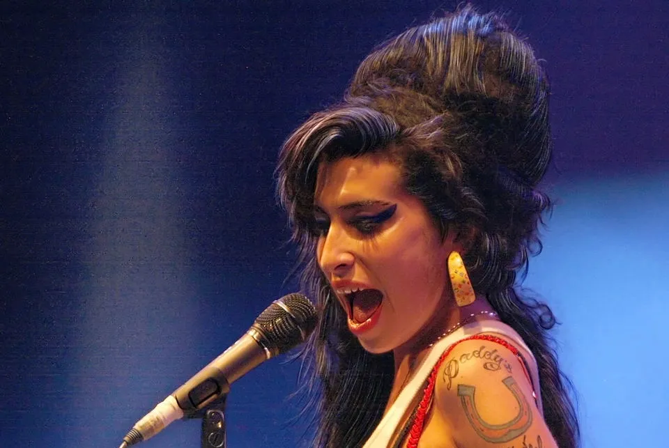 Singer Amy Winehouse performing live on stage with vintage hairstyle and bold makeup, holding a microphone.