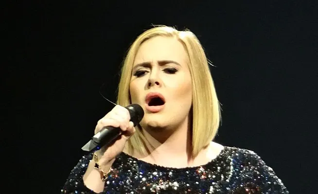 Adele singing passionately on stage, holding a microphone, wearing a sparkling black outfit against a dark background.