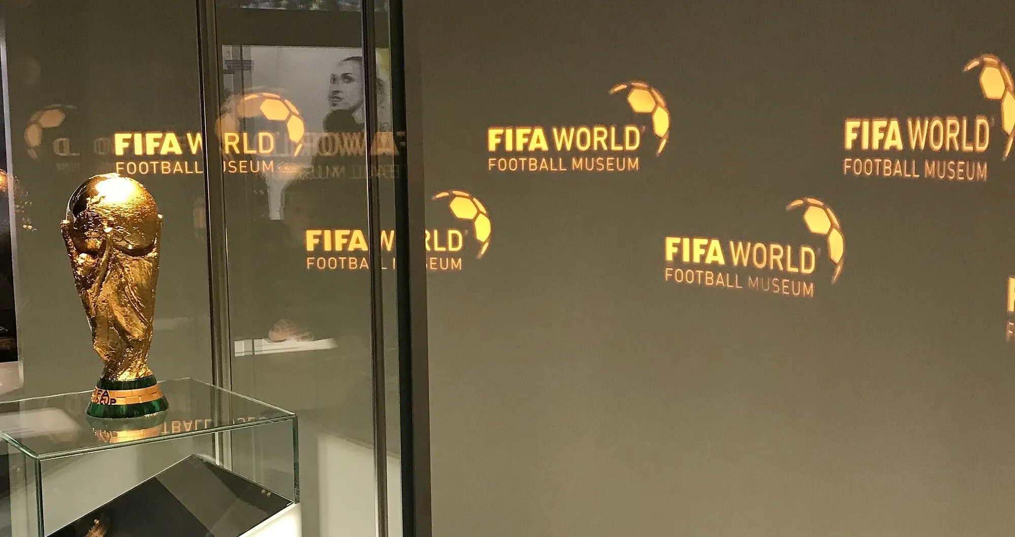 FIFA World Cup trophy displayed at football museum with reflections of FIFA World Football Museum logos on wall.