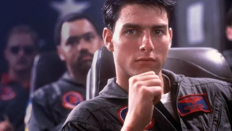 Tom Cruise as a young fighter pilot in the 1980s film, wearing a flight jacket inside a cockpit setting.