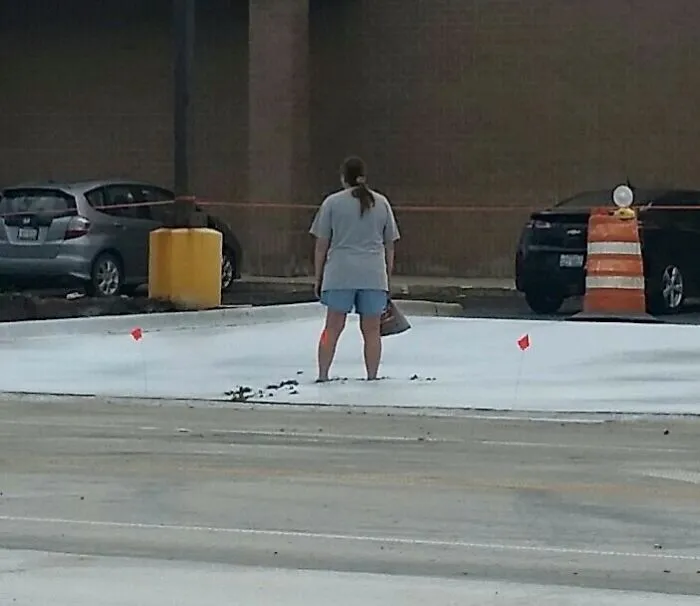 Woman standing in shorts on fresh concrete, surrounded by cars and construction cones, funny photo capturing the moment