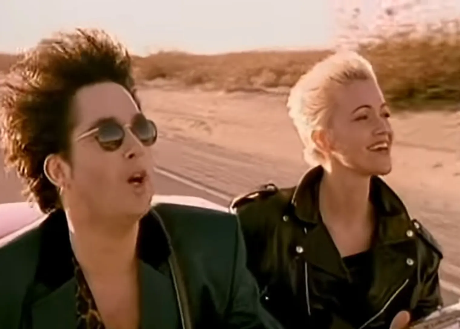 Two band members riding in a convertible outdoors, representing the 1990s band associated with All That She Wants.