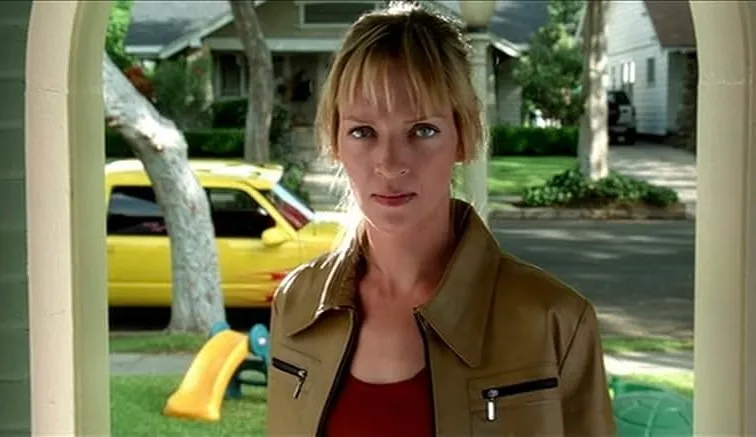 Uma Thurman standing outside a suburban home wearing a brown leather jacket, portraying Buffy in Buffy the Vampire Slayer.
