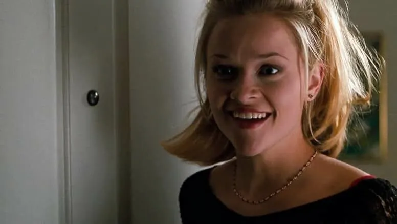 Reese Witherspoon smiling indoors with a black top and necklace in a scene related to Buffy the Vampire Slayer.