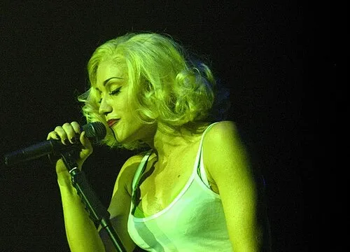 Pop star Gwen Stefani performing on stage with a microphone, wearing a white tank top and blonde hair.