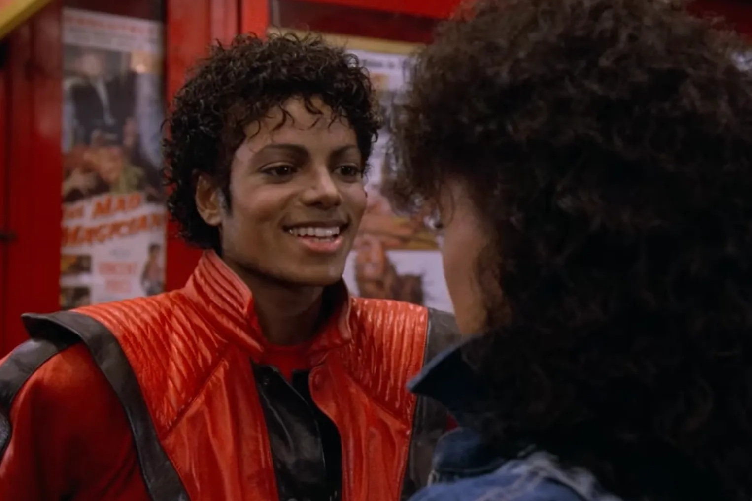 Michael Jackson in the 1980s music video wearing a red jacket, interacting with a woman with curly hair.