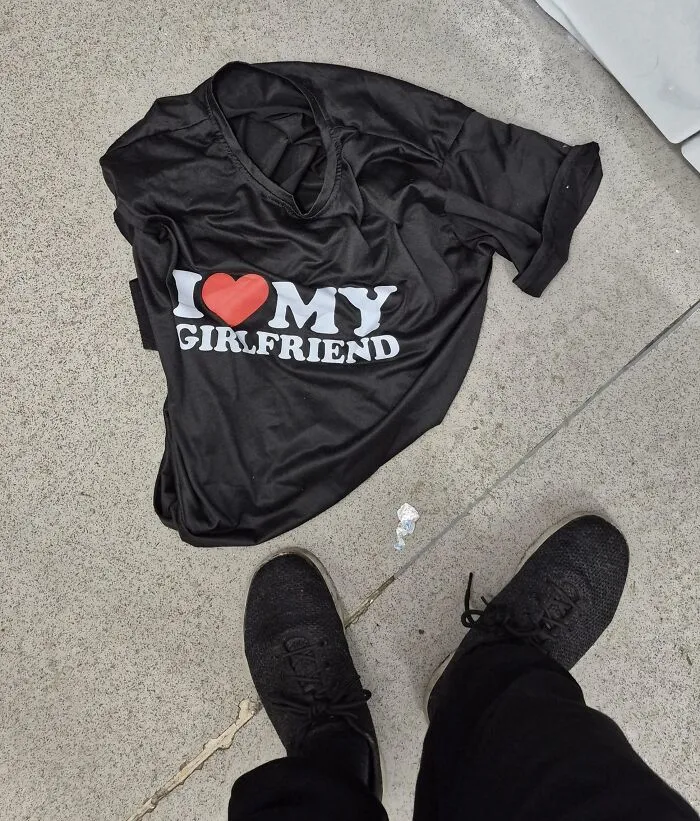 Black T-shirt with I love my girlfriend print lying on concrete floor with black shoes nearby in funny photos context