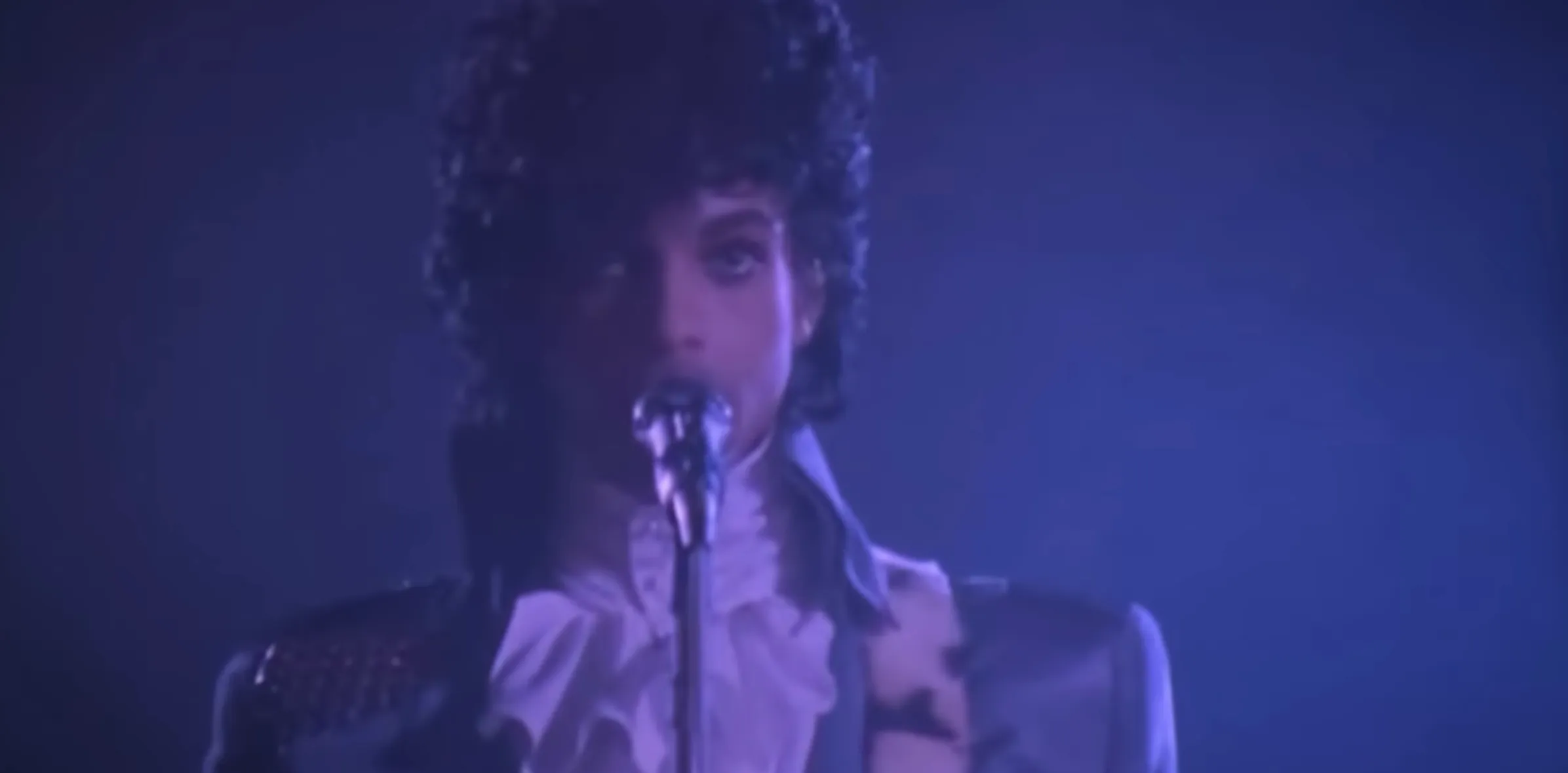 Prince singing into a microphone during the iconic 1980s Purple Rain music video with elaborate choreography.