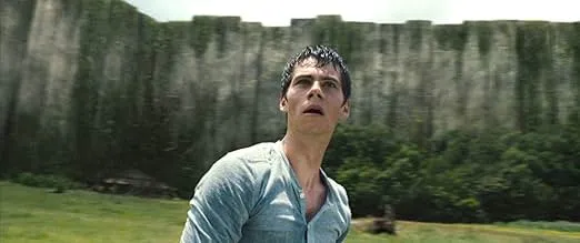Two teenagers in a dystopian reality show setting, facing each other with a forest and wooden structures in the background.