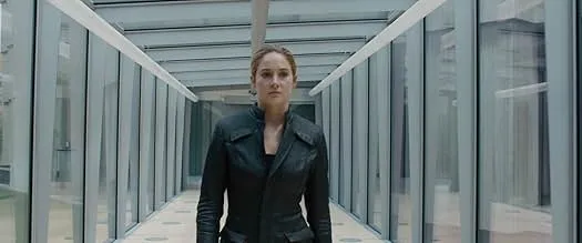 A young woman in a black jacket stands in a futuristic glass corridor, representing the dystopian reality show Divergent.