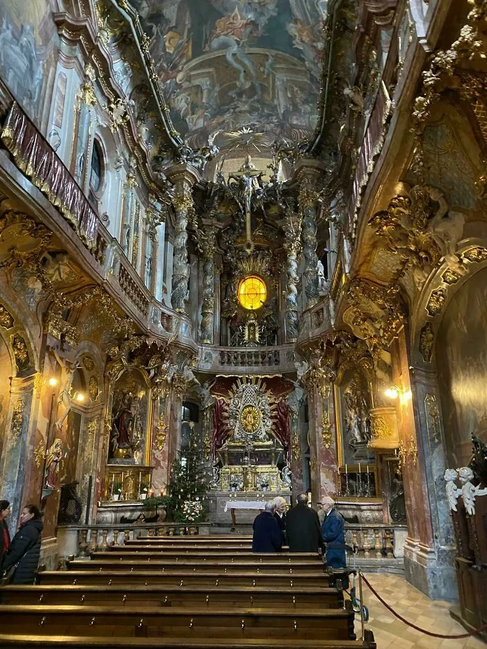 Ornate and beautiful architecture of a church interior with golden decorations, paintings, and people seated in pews.