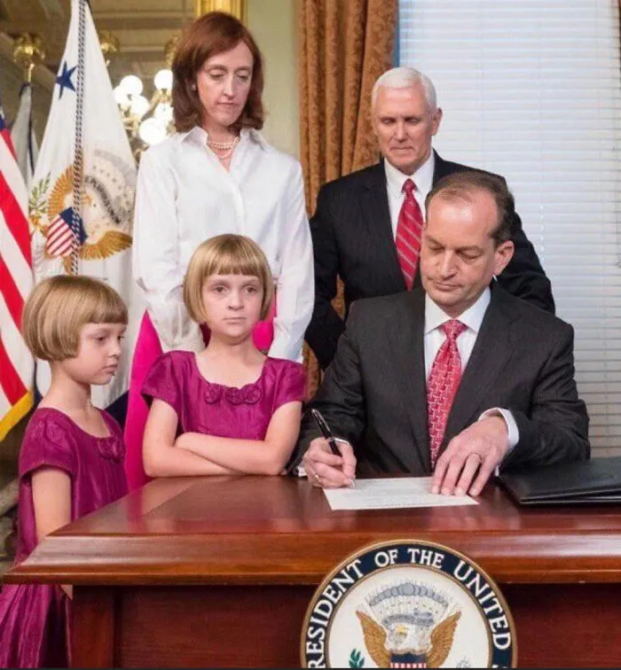 Two young girls with tragic hair accidents stand beside an official signing a document in a formal setting.