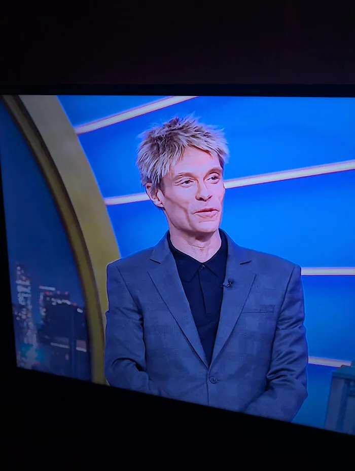 Man with a tragic hair accident hairstyle wearing a gray suit and black shirt on a blue studio background.