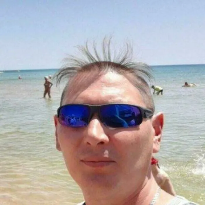 Man at the beach wearing sunglasses with a dramatic, uneven hair accident showing sparse and spiky hair strands.