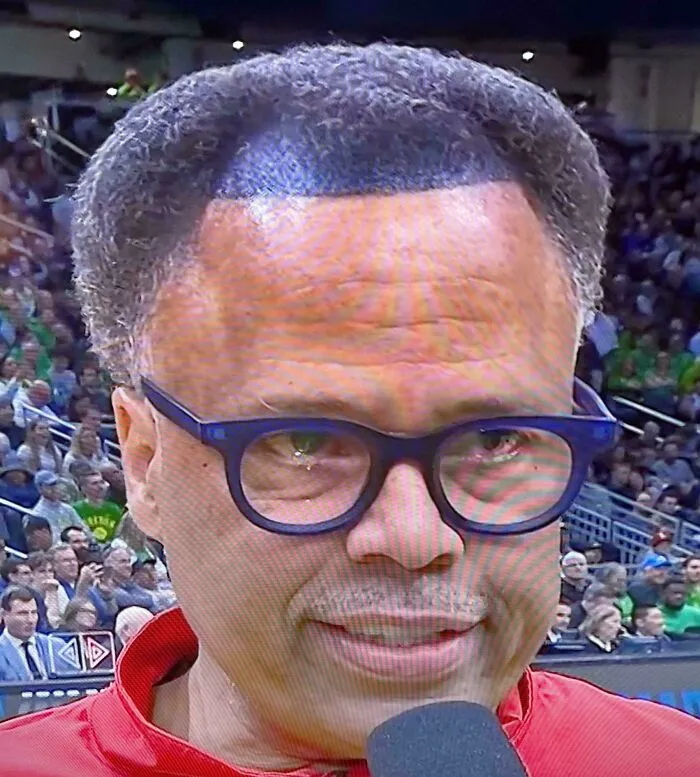 Man with a highly unusual haircut wearing glasses and a red shirt speaking into a microphone at a crowded event.