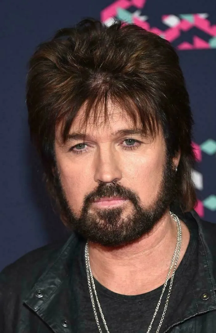 Man with a voluminous 1980s hairstyle and beard wearing a black leather jacket and silver chains at an event.
