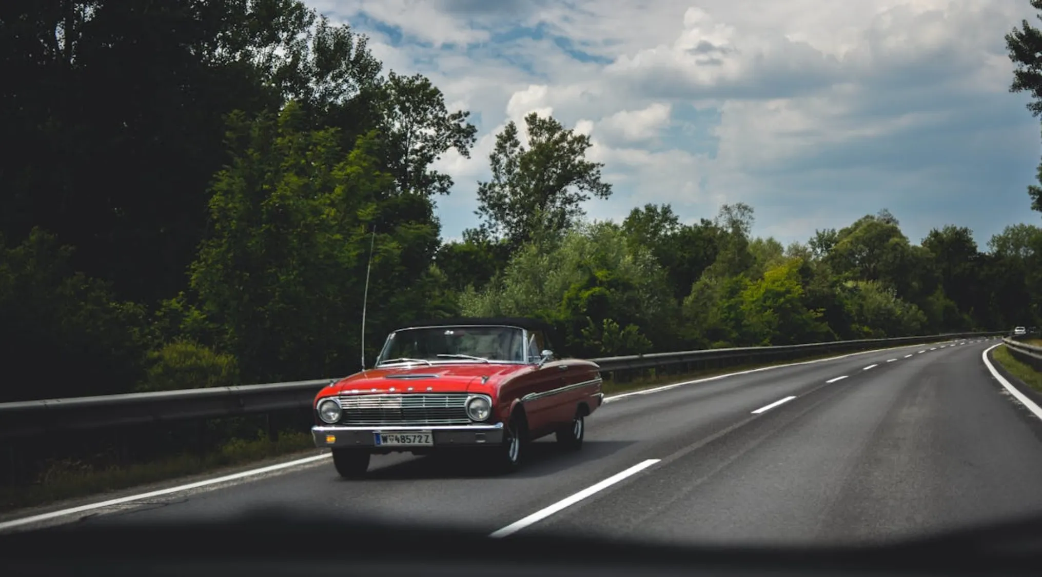 A red vintage car driving on a highway surrounded by trees under a partly cloudy sky showing basic math skills.
