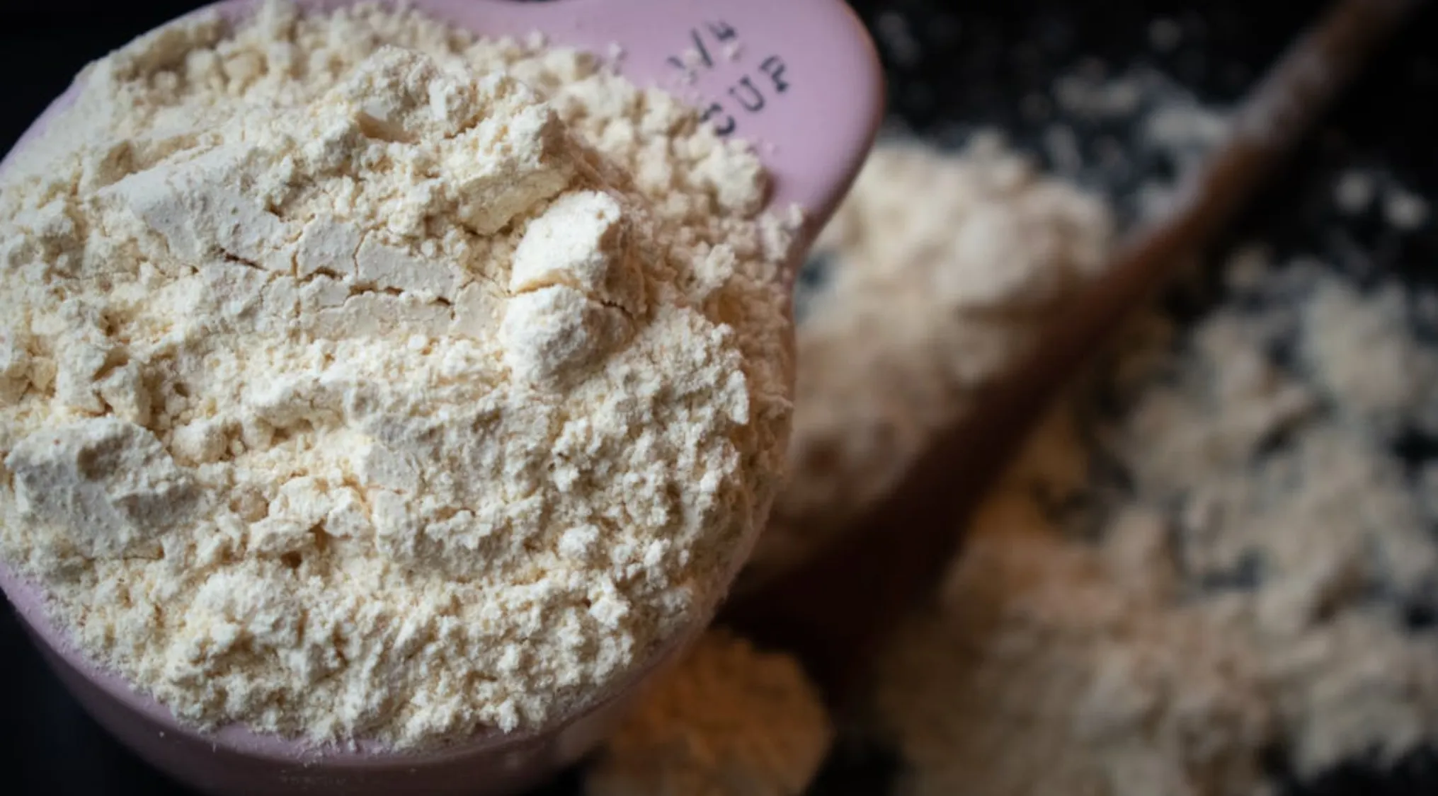 Measuring two-thirds cup of flour with a pink measuring spoon on a wooden surface with scattered flour.