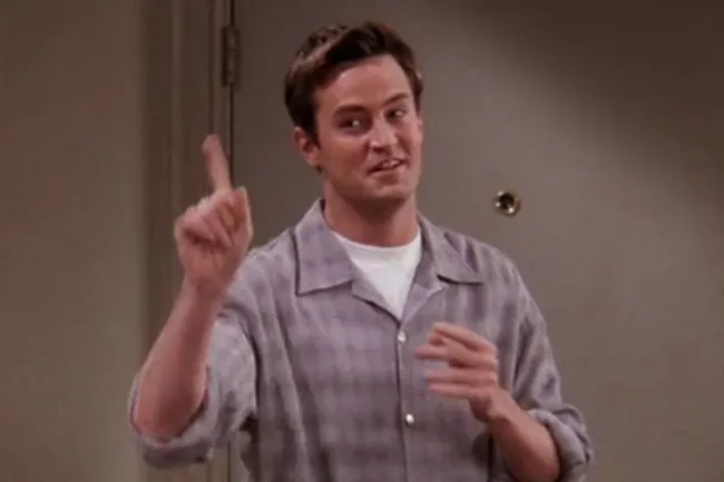 Chandler Bing from old sitcom Friends smiling and gesturing with finger raised indoors in casual shirt.