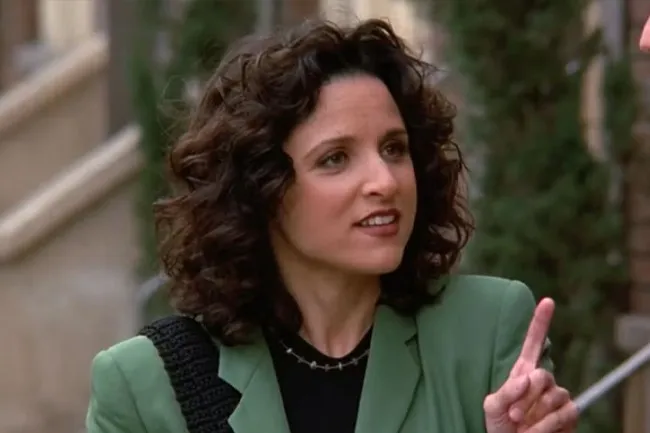 Elaine Benes from old sitcom, wearing green jacket, curly hair, gesturing with finger, outdoors background