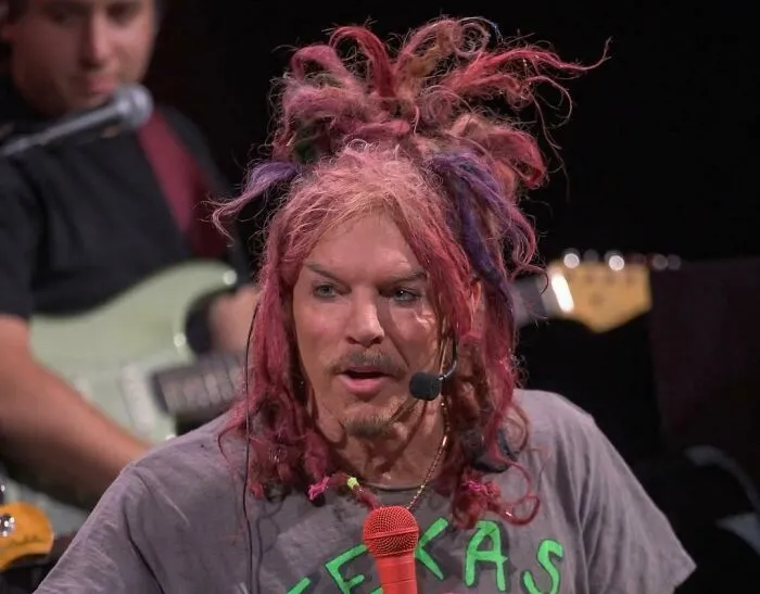 Man with messy, multicolored hair styled chaotically, speaking into a microphone during a live music performance.