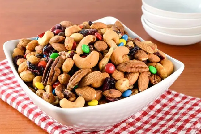 Bowl of colorful trail mix with nuts, dried fruit, and candy pieces on a red checkered cloth.