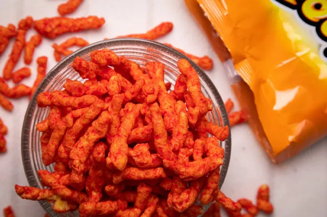 Bowl filled with spicy Hot Cheetos snack pieces next to an opened bright orange bag on a white surface.