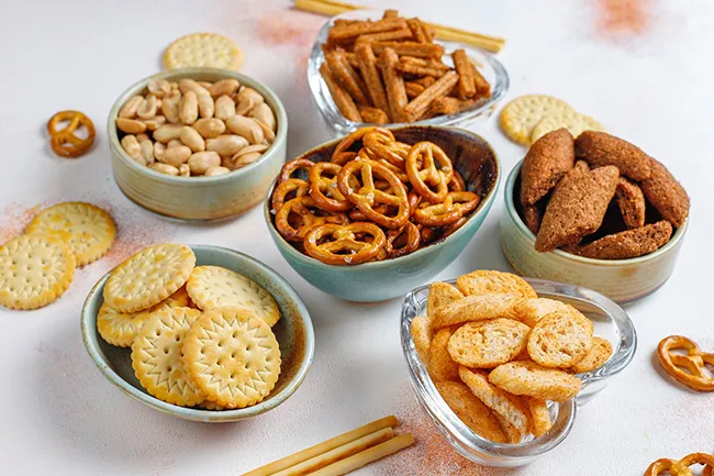 Assorted nostalgic snacks including pretzels, crackers, peanuts, and various savory treats in bowls on a white surface.