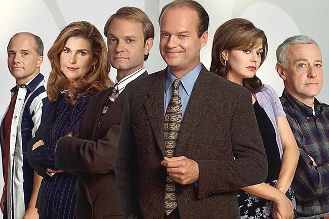 Cast of the comfort TV rerun Frasier standing together in character, dressed in 90s style business and casual attire.