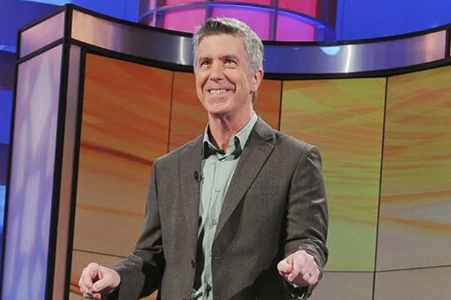 Host on America's Funniest Home Videos set, smiling and wearing a gray suit with colorful studio background behind him.
