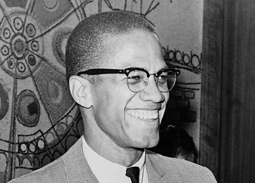 Black and white photo of Malcolm X smiling, wearing glasses and a suit, relating to I Have a Dream speech and New York.