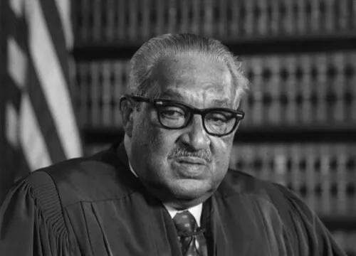 Thurgood Marshall wearing glasses and judicial robe, seated in front of bookshelves, representing Washington, D.C.