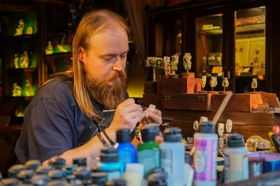 Artist preparing to paint a figure carefully, surrounded by paint bottles and miniature sculpted figures in a detailed workspace.