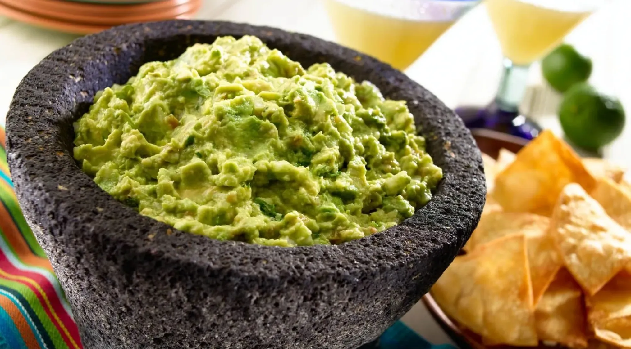 Close-up of fresh guacamole made with avocado in a stone mortar served with tortilla chips and lime in the background.