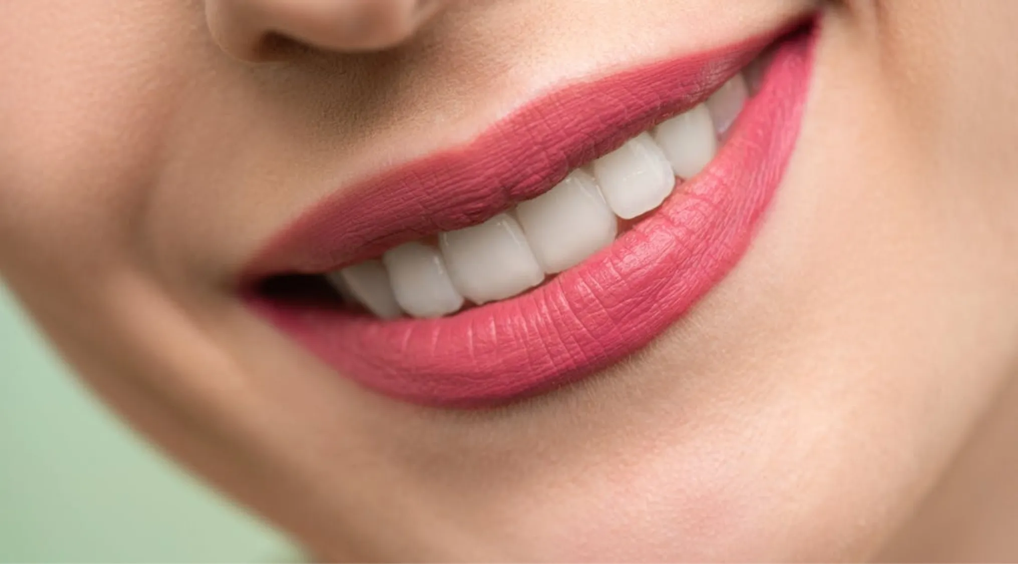 Close-up of smiling lips with pink lipstick showing clean white adult human teeth in a natural skin tone background.