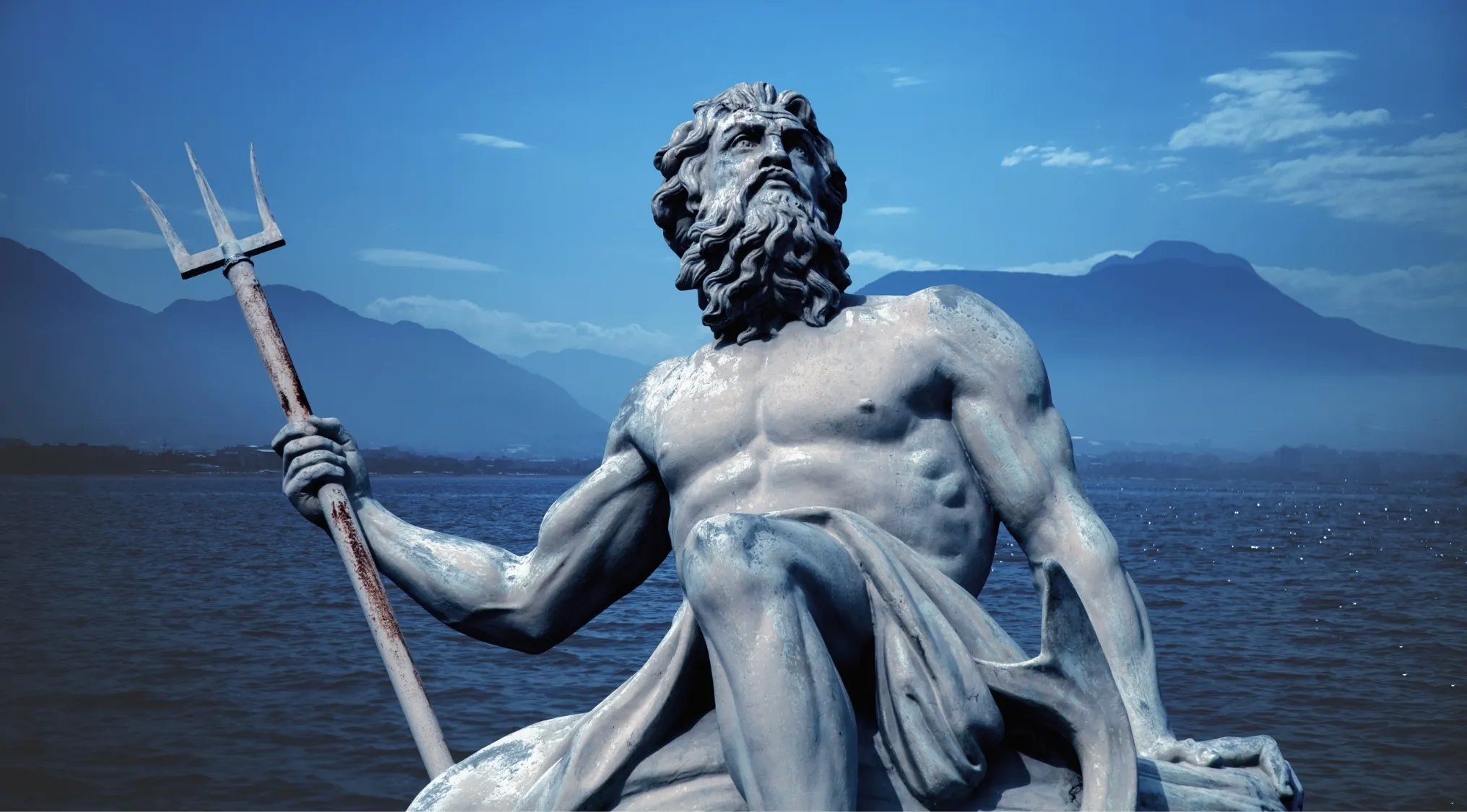 Statue of the Greek god of the sea holding a trident with ocean and mountains in the background on a clear day.