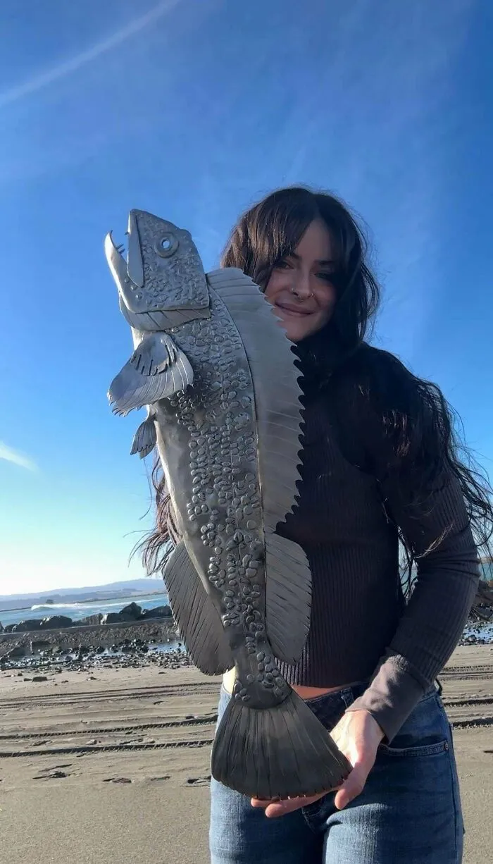 Woman holding a large detailed metal fish sculpture on a beach, showcasing remarkable metalworker craftsmanship.