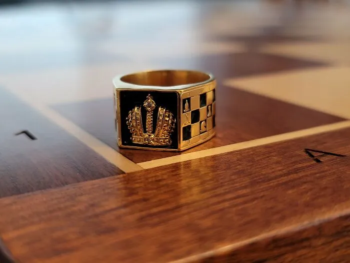 Gold ring with a detailed crown and chessboard pattern, showcasing metalworkers' skill and creativity on a wooden chessboard.