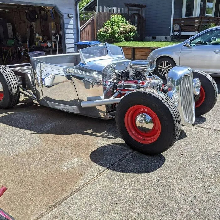 Custom polished metal hot rod with exposed engine and red wheels parked outside a garage showcasing metalworkers' craftsmanship.