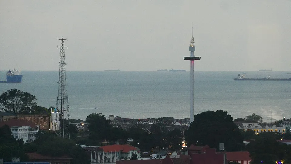 View of the Strait of Malacca with ships passing between two landmasses at dusk, urban buildings in foreground.