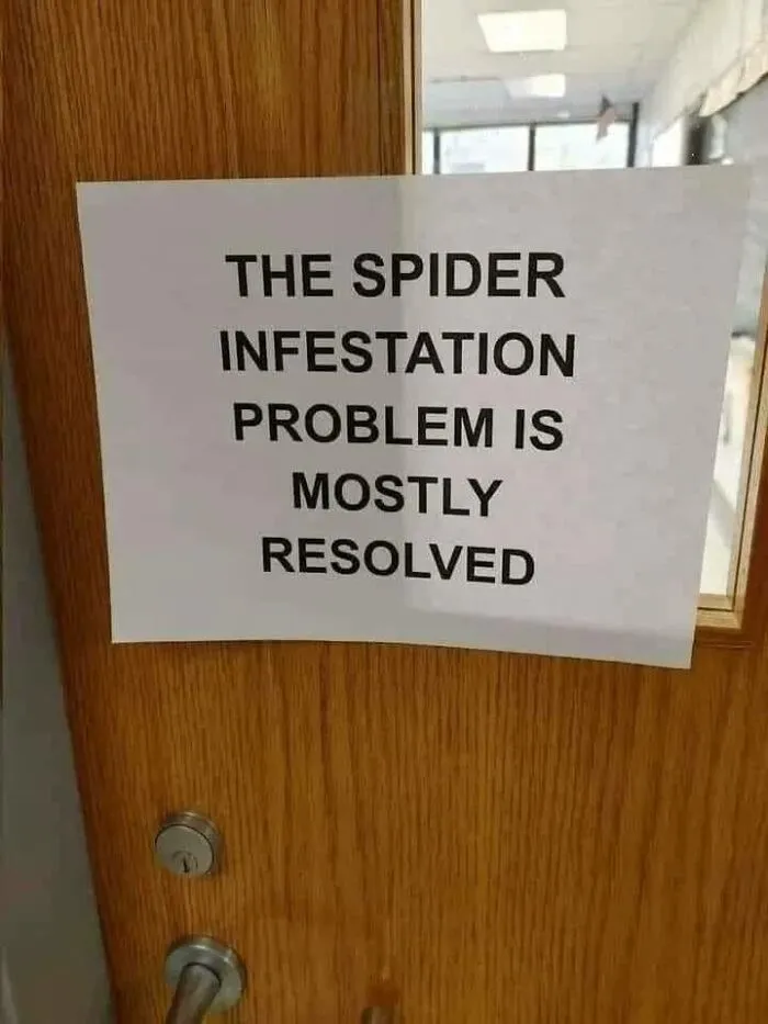 Sign on wooden door humorously stating the spider infestation problem is mostly resolved for double take moments.