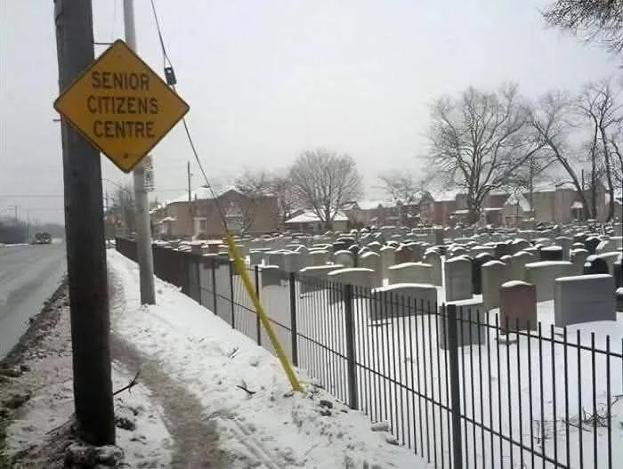 Unusual street sign for senior citizens centre placed next to a snowy cemetery, creating a strange and ironic scene.