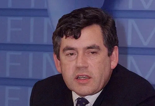 British political figure Gordon Brown speaking at a formal event, wearing a dark suit and tie.
