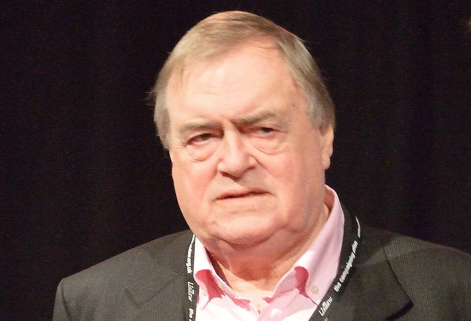 John Prescott in a black suit and pink shirt speaking at an event against a black background.