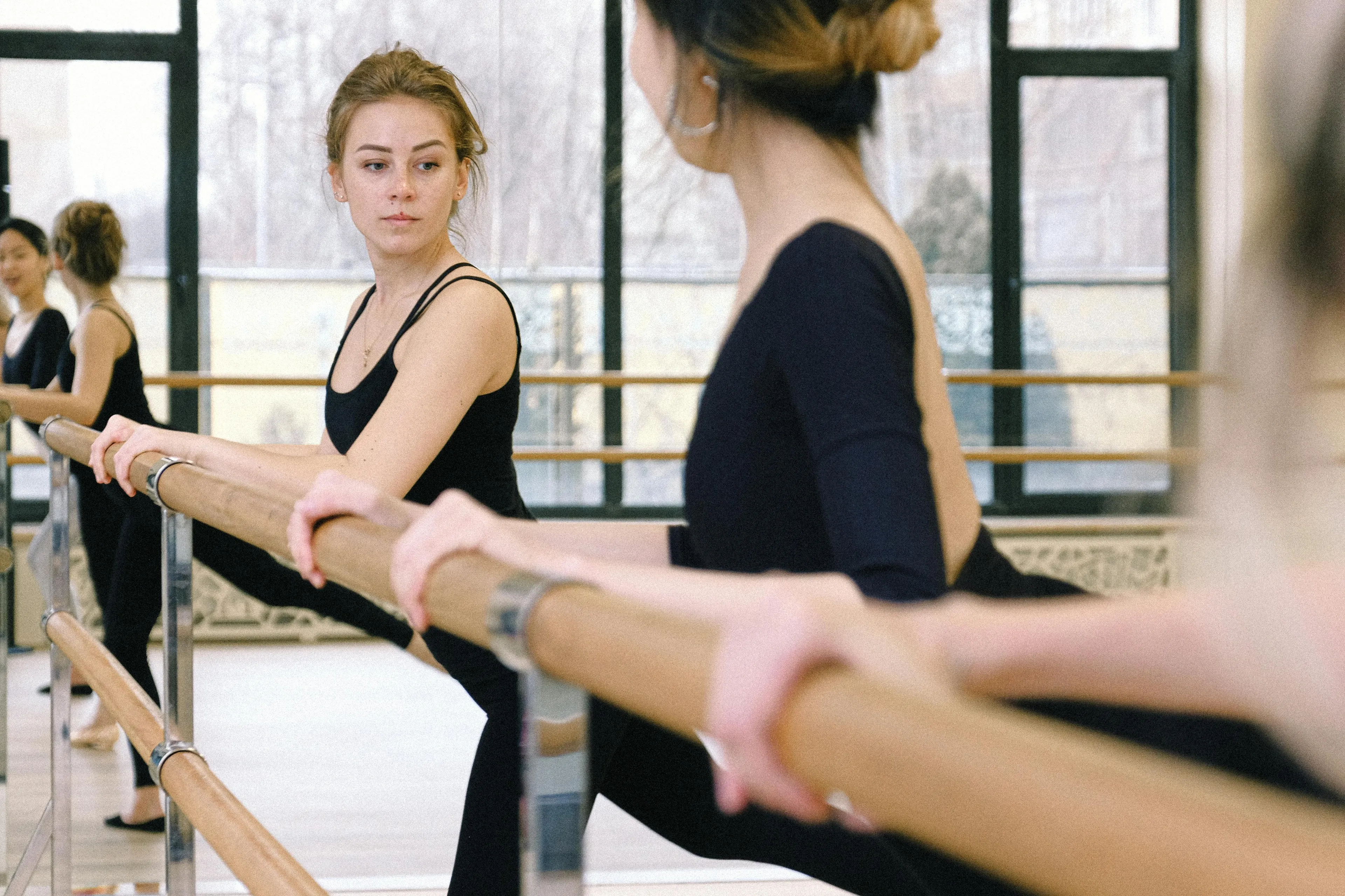 Women in black workout clothing using a barre in a dance studio for balance and support during training.