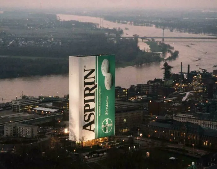 Massive aspirin billboard towering over industrial cityscape at dusk, illustrating dystopian urban hell and modern life challenges.