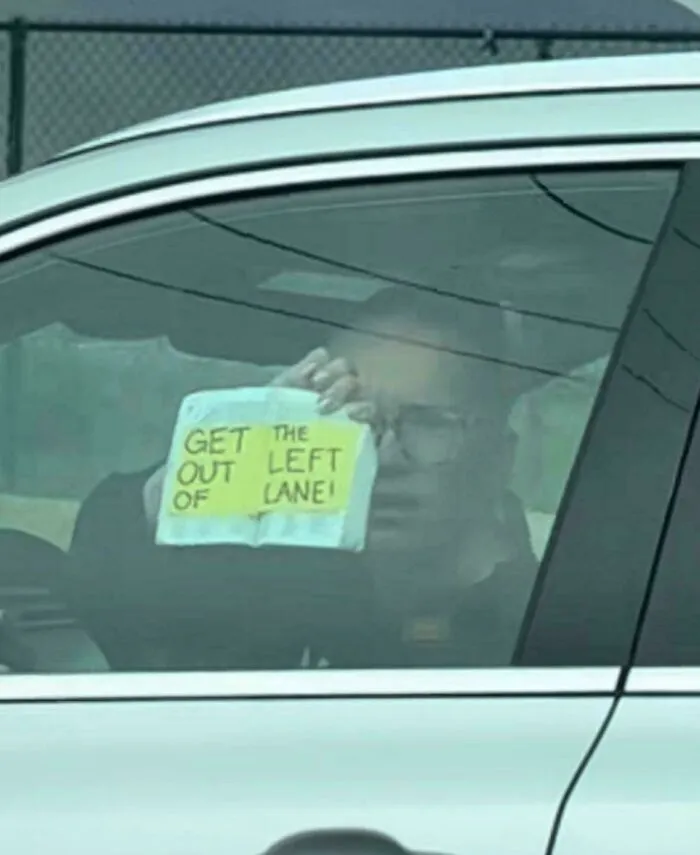 Man inside car holding a handwritten sign saying get out of the left lane, a comedic sign that went wrong.